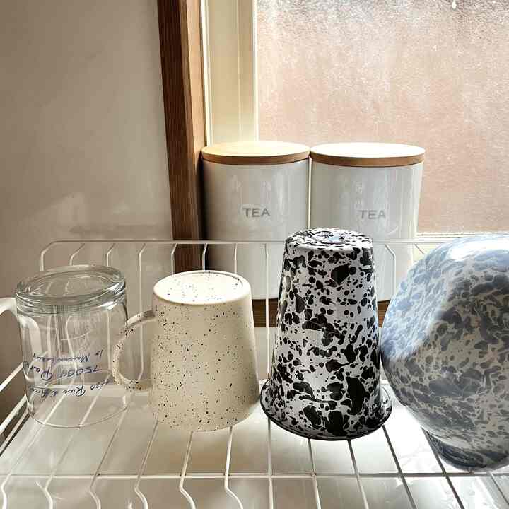 白とナチュラルカラーのキッチン、窓辺の食器乾燥ラックに並んだ様々なマグカップとボウルが印象的なインテリア