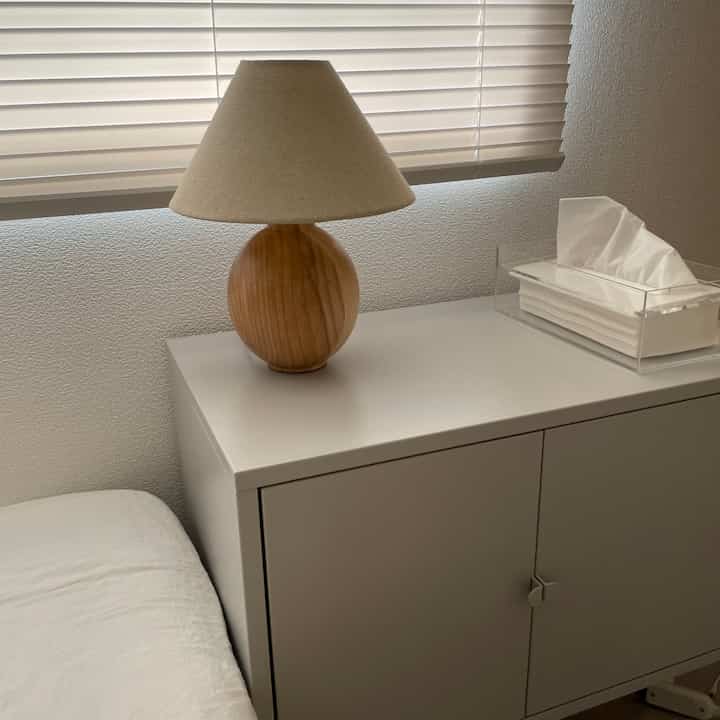 White-toned monotone bedroom featuring a white cabinet as nightstand with wood-tone lamp and white blinds, creating a neat atmosphere