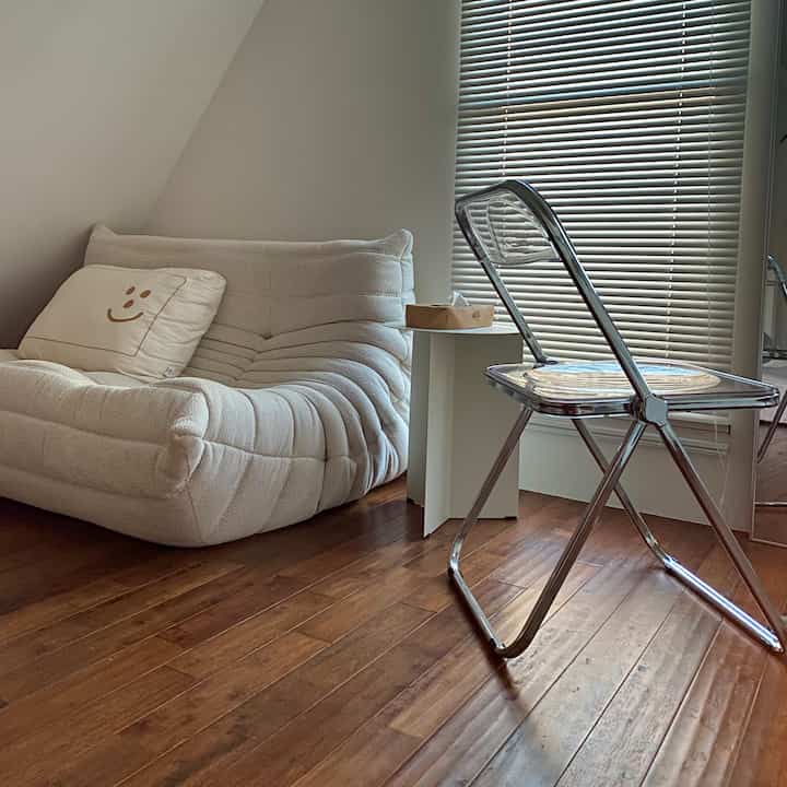 White and brown toned living room featuring soft sofa, transparent chair, and blinds creating a cozy atmosphere