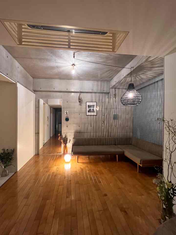 グレーのコンクリート壁と天井、ブラウンの木製床、ローバックのコーナーソファが配置されたナチュラルなワンルームのリビングルーム