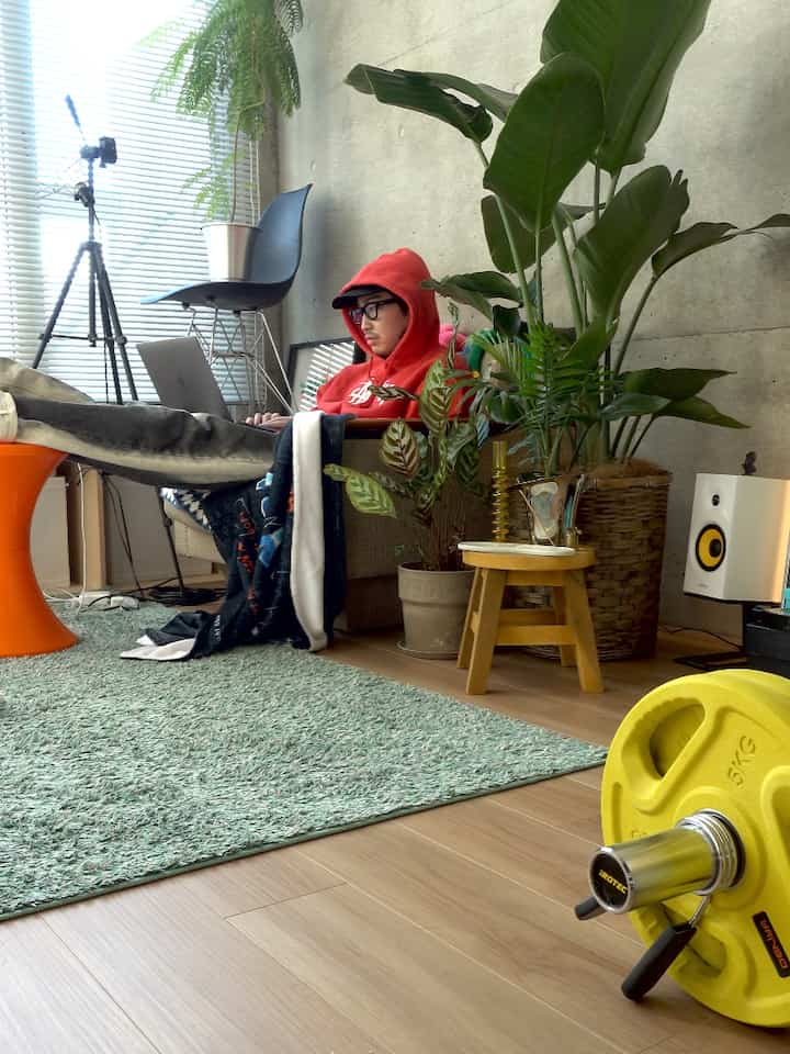 グレーのコンクリート壁と木目調の床のリビング、ソファと植物が配置され、ノートパソコンでテレワーク中の空間