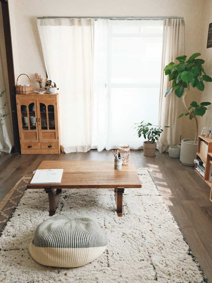 ライトブラウンの木目調フロアと家具が配置されたナチュラルモダンなリビング、ホワイトカーテンと柔らかなフロアクッションがある雰囲気の良い空間