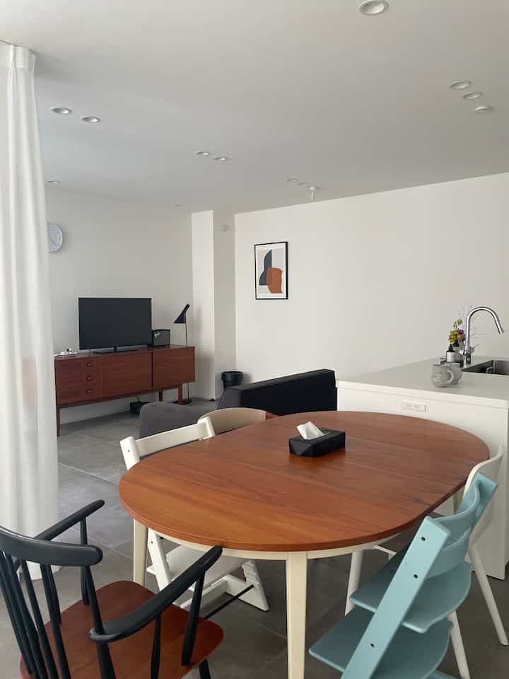 Modern natural living and dining space with white walls, gray tiled floor, warm wooden dining table, and floor lamp
