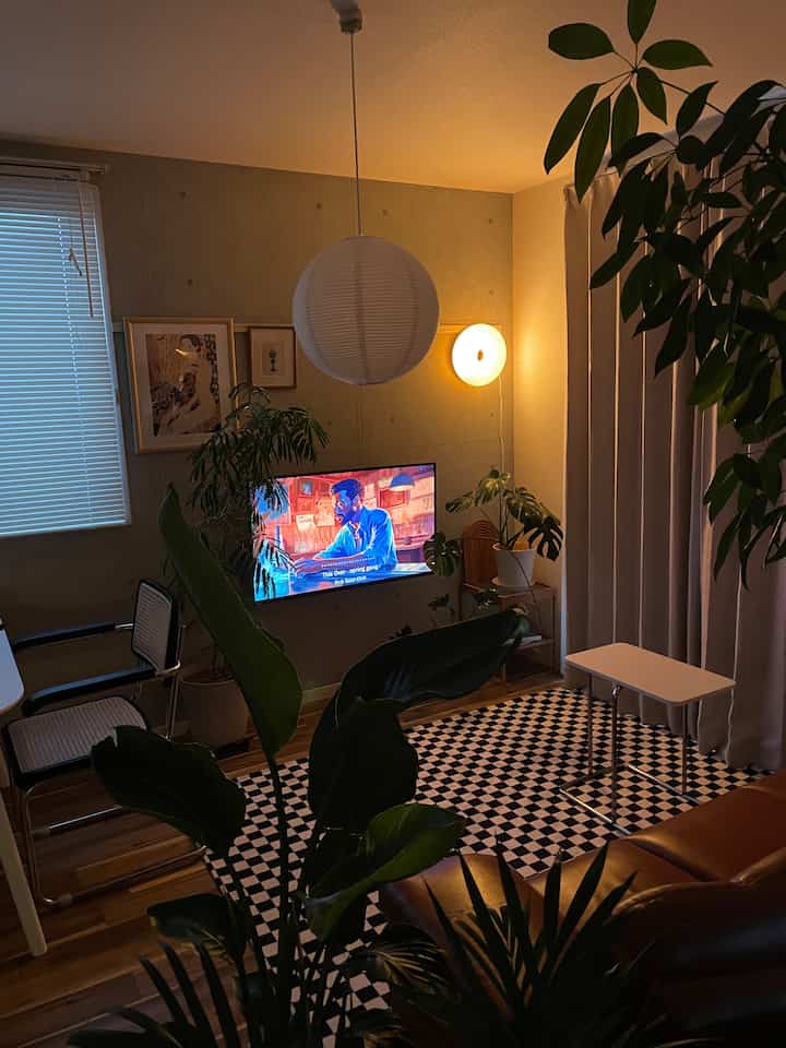 Dark-toned modern living room featuring concrete wallpaper, large plants, checkerboard rug, and cozy lighting