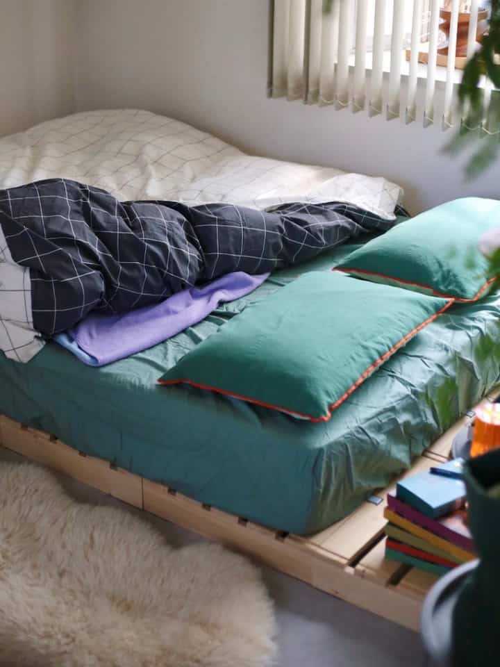 ベージュの壁と木目調のベッドが配置されたコンパクトな寝室、グリーンの寝具と枕がシンプルモダンな雰囲気を演出するインテリア