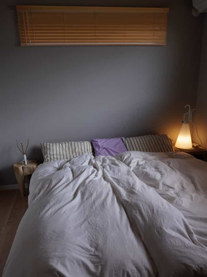 グレーの壁と木目調ブラインド、白い寝具が配置された寝室、ディフューザーが置かれた雰囲気のある空間