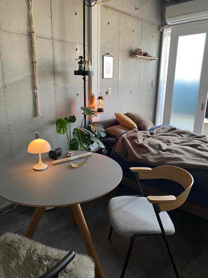 グレーのコンクリート壁とナチュラルウッド家具が調和した一人暮らし用寝室、温かみのあるテーブルランプと居心地の良いアームチェアが配置された印象的な空間