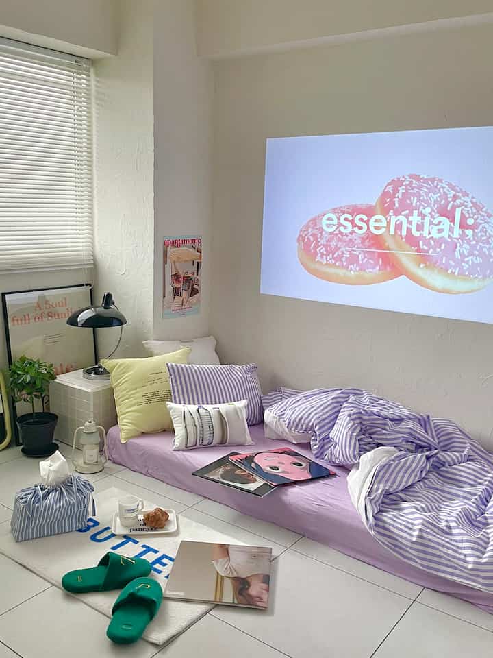A small bedroom in white and lavender tones featuring a floor mattress with cushions and a projected image on the wall, creating a cozy atmosphere