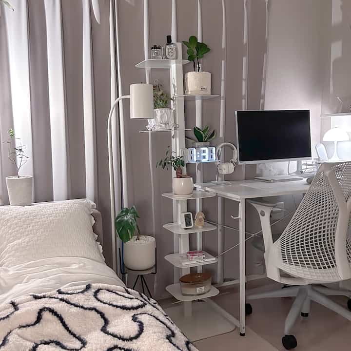 Bright white-toned compact bedroom and home office with bookshelf and various plants creating a cozy atmosphere
