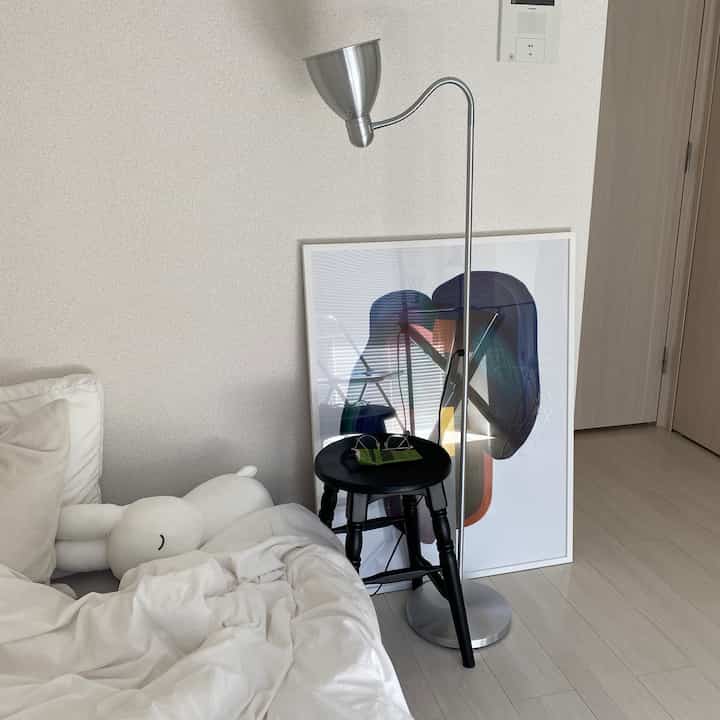 白を基調とした寝室、ベッドとフロアランプ、黒いスツールとアートが配置されたシンプルでナチュラルな印象のインテリア