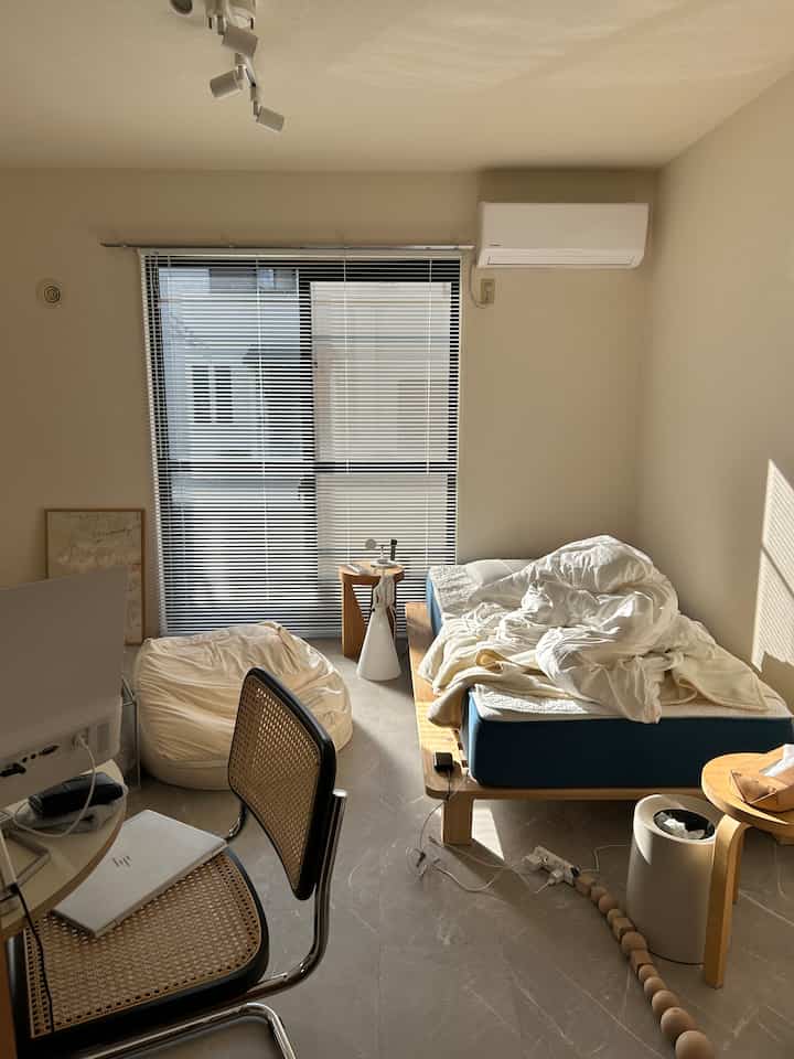 自然光が差し込むナチュラルカラーと白を基調としたワンルーム寝室、シングルベッドとビーズクッション、作業スペースが配置された快適な空間