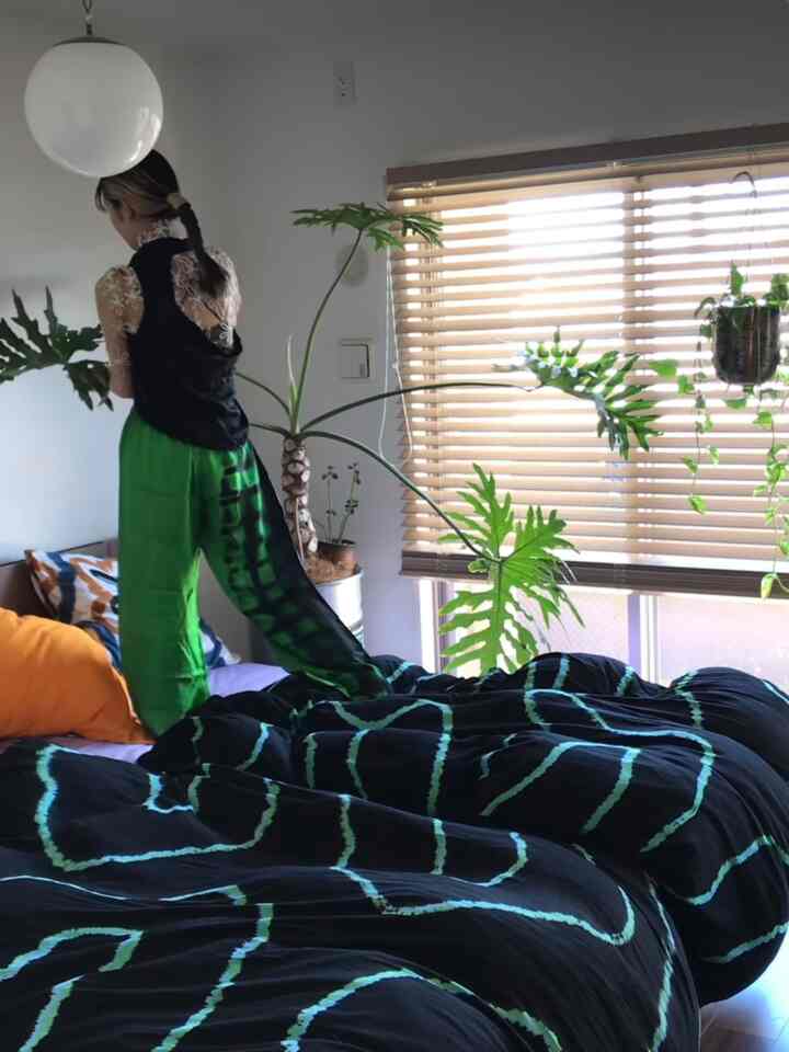 白い壁と窓のブラインドがある寝室、緑の植物と黒地に青緑の模様の寝具が配置された雰囲気のある空間