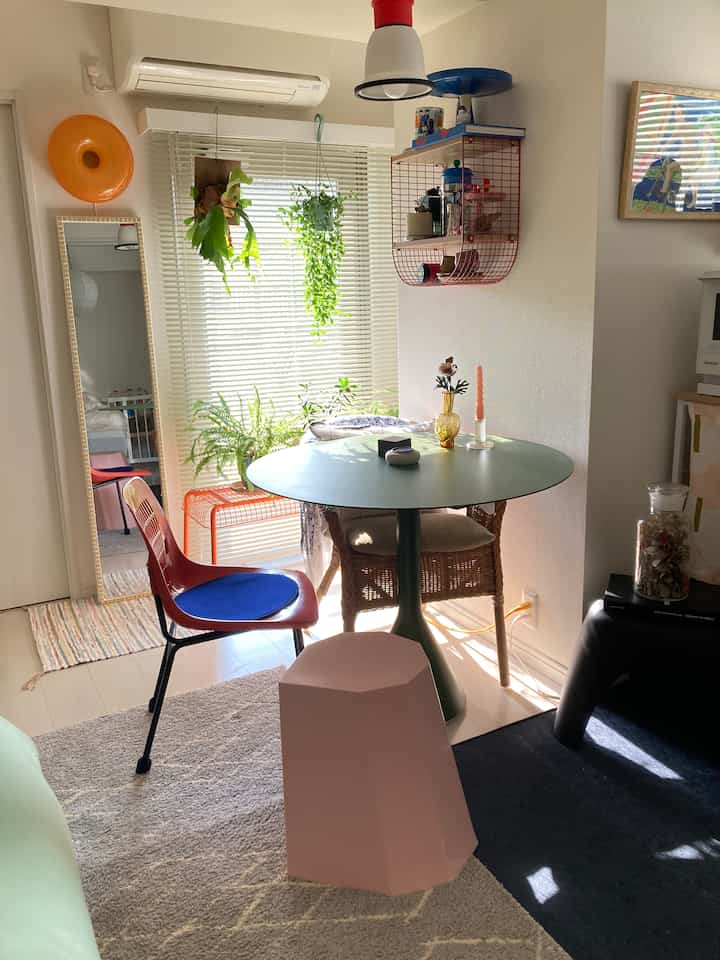 A 1LDK living room in white and natural tones featuring a round dining table, diverse chairs, and cozy mid-century modern decor