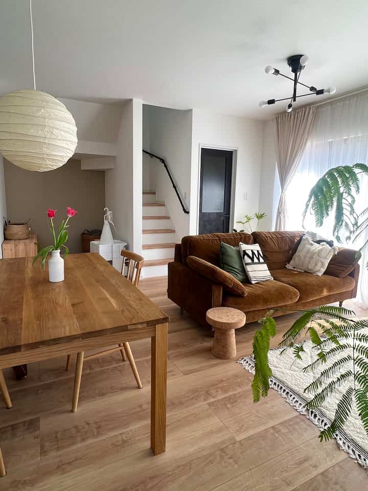 White and brown toned living-dining space featuring corduroy sofa, wooden dining table, and large plants with warm, natural ambiance