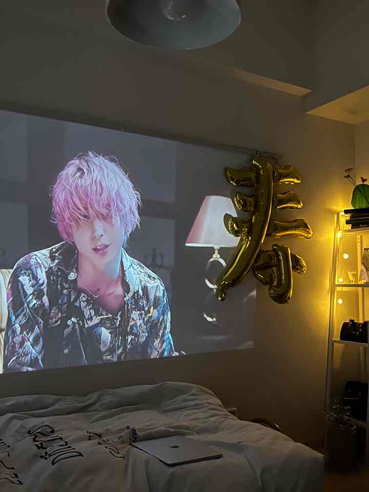 白を基調とした寝室、壁にプロジェクター画面が映し出され、ゴールドのバルーン飾りと棚、ベッドが配置された印象的な空間