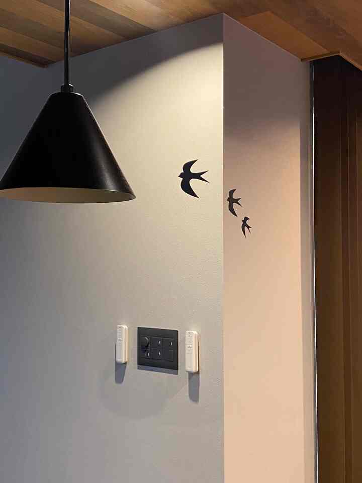 白い壁とゴールドの光が醸し出す玄関、壁に飛ぶ鳥のステッカーと黒いペンダントライトが配置された空間