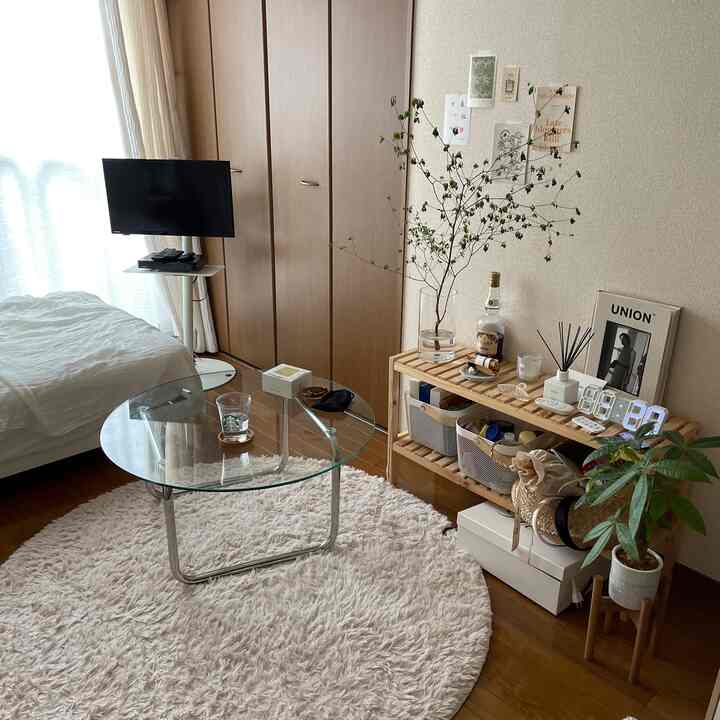 ベージュ色の壁と木目調の床がある寝室、丸いガラステーブルとテレビ台が配置された自然でシンプルなインテリア