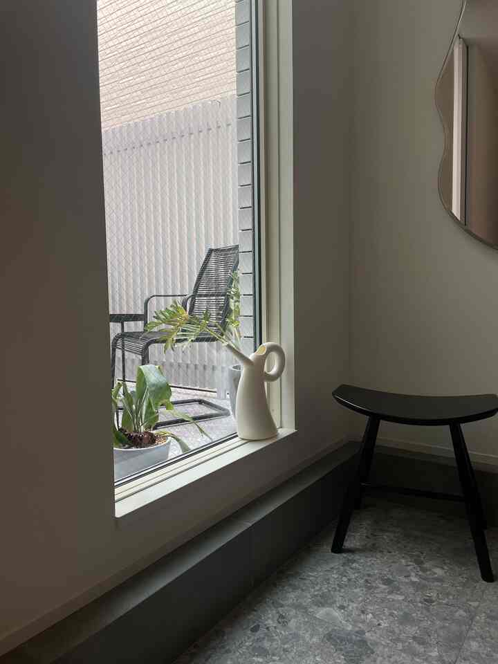 A simple entrance space with white and black tones, featuring a stool and plants beside a window overlooking a balcony with chairs