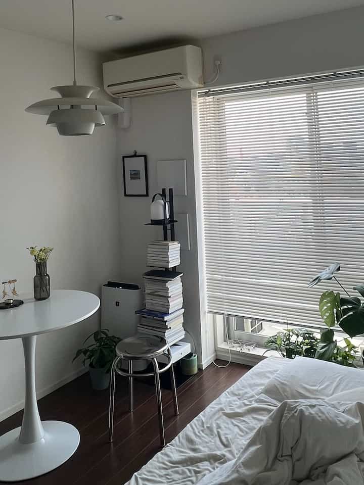 White and dark brown toned studio bedroom with simple interior, featuring window blinds, a stool, and a round dining table near the window, creating a cozy atmosphere