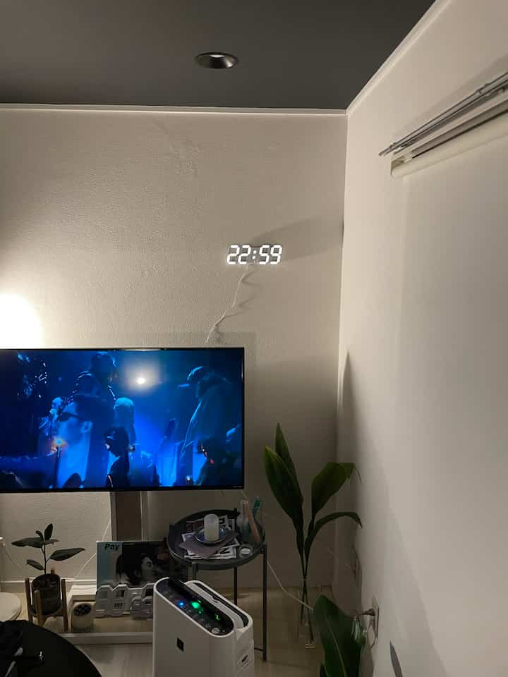 A white-toned living room featuring TV stand, side table, and an LED digital clock on the wall with cozy indirect lighting