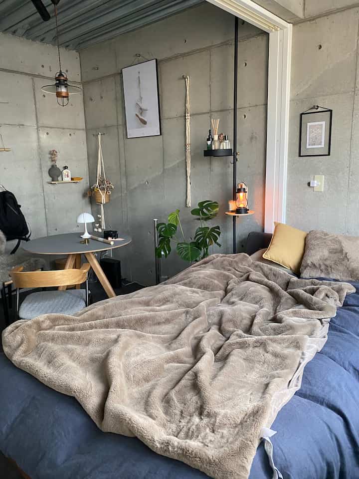 グレーのコンクリート壁とネイビーのベッド、ベージュのブランケットが配置された雰囲気のある寝室