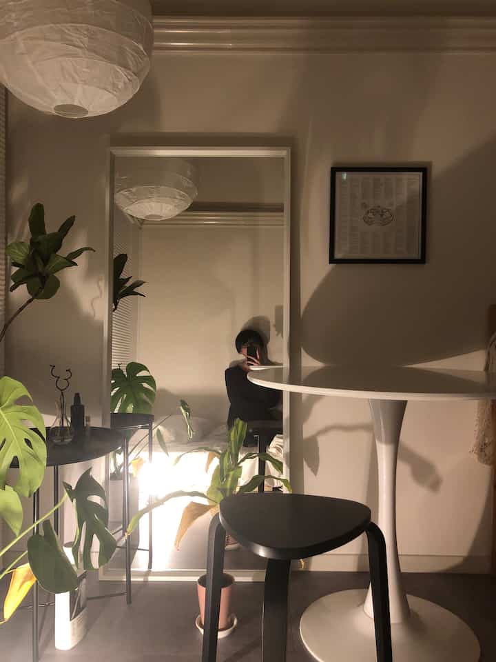 White and green toned studio apartment featuring a large mirror, white round table, black stool, and plants creating a simple natural space