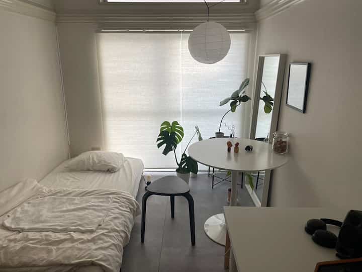 Small white and gray toned room featuring a round dining table, stool, bed, and plants creating a simple natural atmosphere