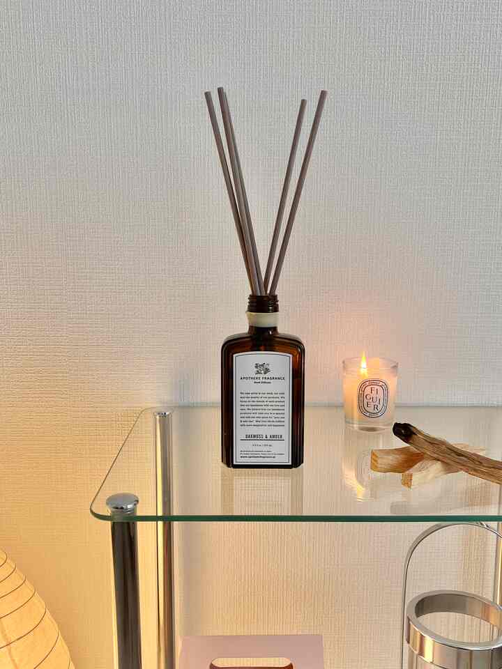 透明なガラスラックの上に茶色のディフューザーとキャンドルが置かれた、温かみのあるモダンな寝室のインテリア