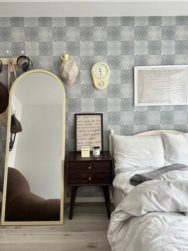 パターン壁紙のあるナチュラルなブラウン系の寝室、白い寝具と大型のフロアミラー、ヴィンテージ風の黄色い壁時計が調和するシンプルな空間
