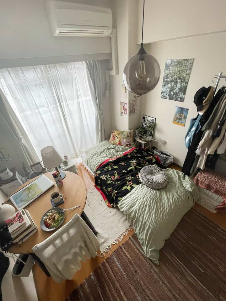 Natural brown-tone studio bedroom featuring a small round dining table, clean curtains, cozy cushions, and warm lighting