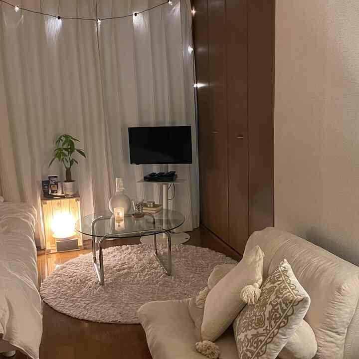 白と茶色のトーンのリビング兼寝室空間、透明なガラスのテーブルとラグ、ソファとカーテンが配置されたシンプルなインテリア