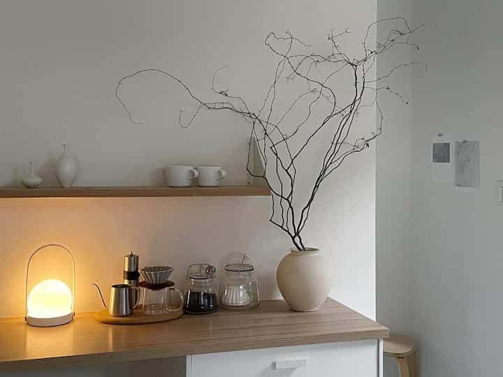 Simple home cafe space in white and wood tones featuring coffee gear, table lamp, and large decorative objet with minimal design