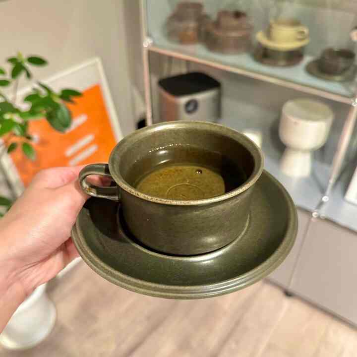 Natural green toned Nordic vintage teacup and saucer held in hand with a glass-front cabinet displaying tableware in the background