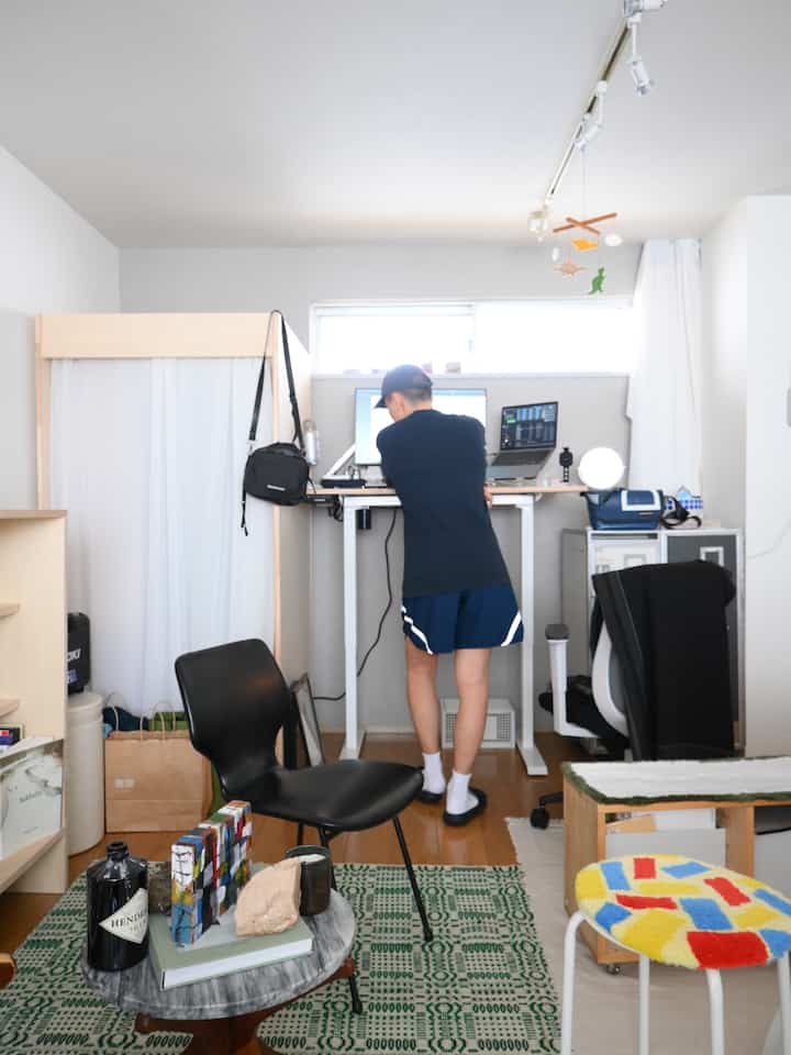 白い壁と茶色い木製床のワンルームのホームオフィス、電動昇降デスクと椅子、カラフルなスツールが配置された明るい作業空間