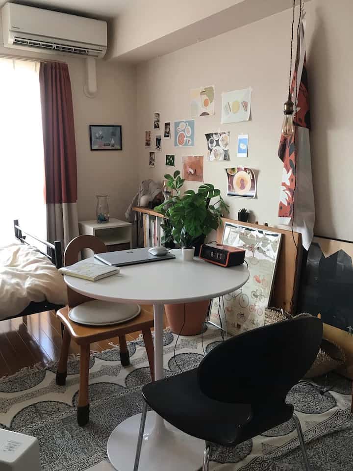 White and brown toned studio bedroom featuring a round dining table, chairs, and a Monstera artificial plant in a cozy modern self-care space