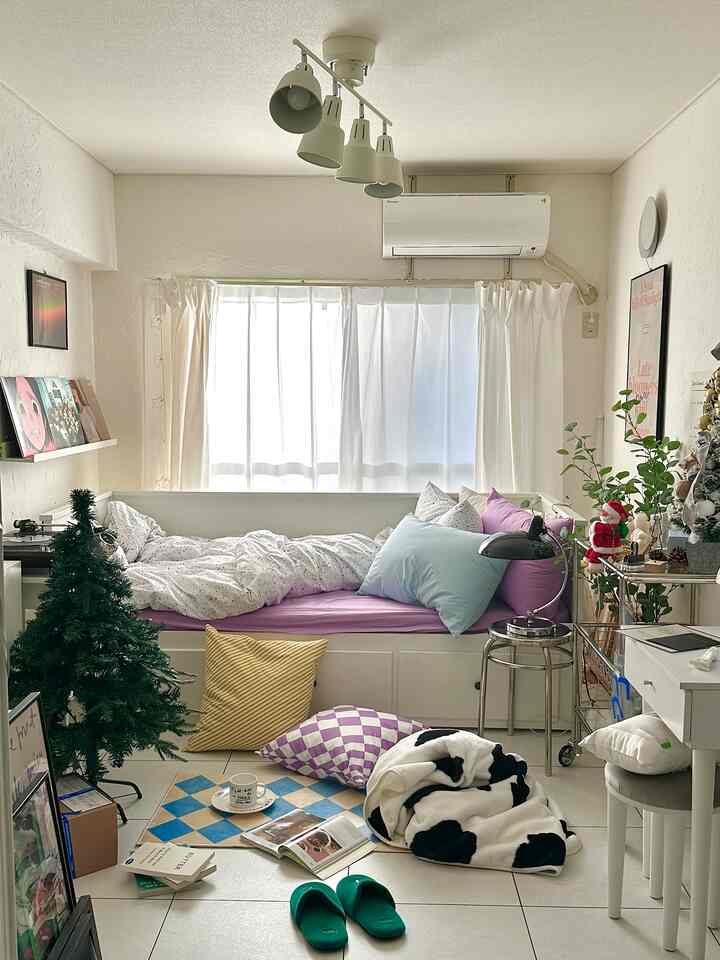 ホワイトとパープルの色調の寝室、窓辺に配置されたベッドとクッション、多彩なブランケットと床に置かれた本がある雰囲気のある空間