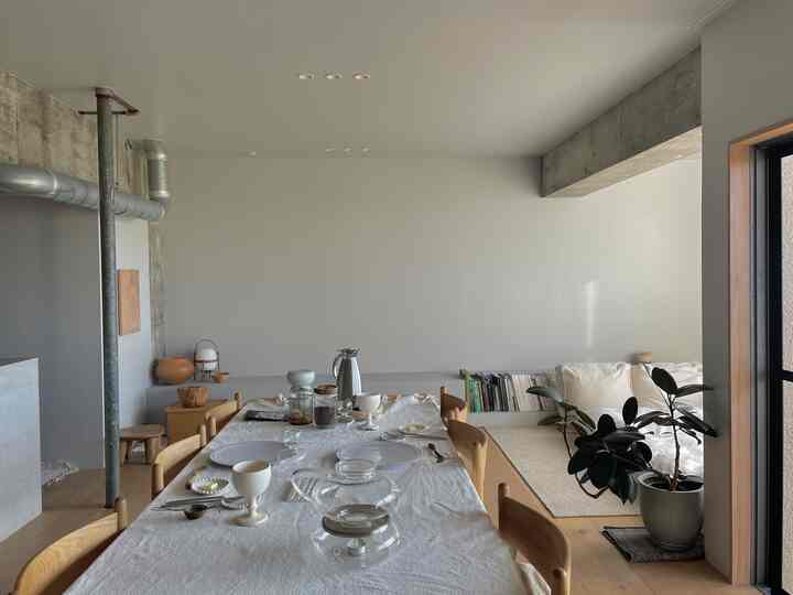 ナチュラルトーンのダイニングルーム、木製テーブルと食器が配置され、白い壁とカーテンが調和した落ち着いた空間