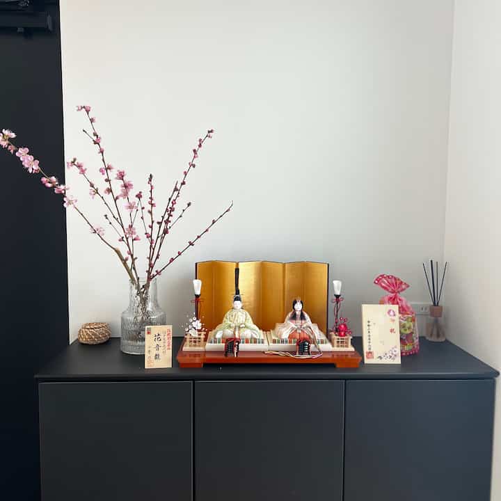 A traditional display featuring hina dolls and a clear vase with pink peach blossoms on a black console against white walls