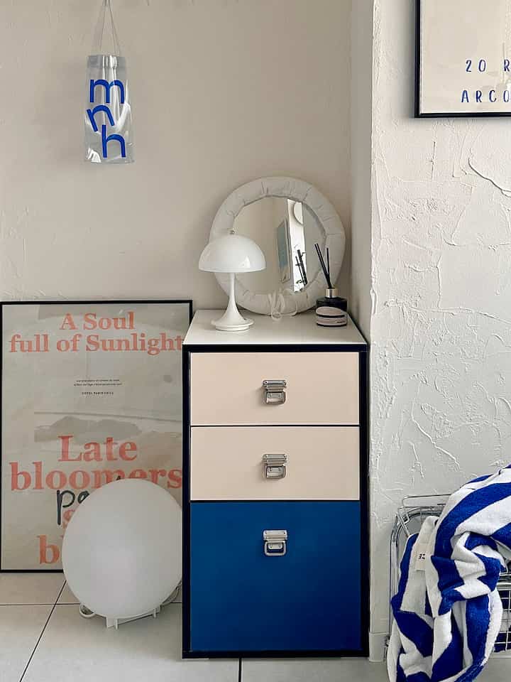 白い壁と青のアクセントが調和するコンパクトな空間にチェスト、円形のファブリックミラー、テーブルランプが配置された雰囲気のあるナチュラルモダンなインテリア