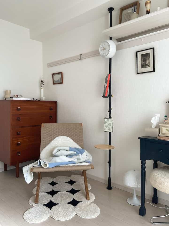 白い壁と明るい床色のナチュラルな雰囲気の狭い部屋、木製のチェストと編まれたアームチェア、模様のあるラグが配置された印象的な空間