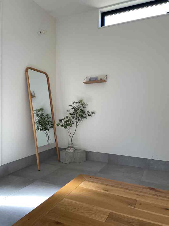 白とグレーの背景に木目調の鏡と棚、緑の植物が配置されたシンプルで自然な雰囲気の玄関スペース