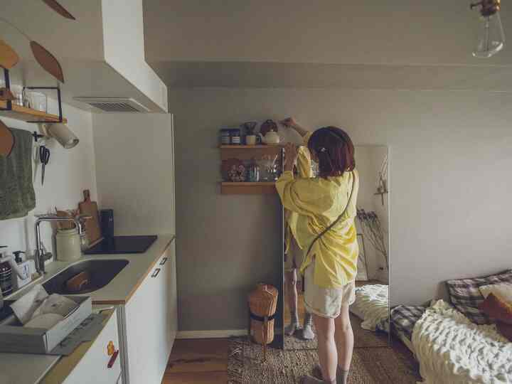 ナチュラルカラーのワンルームのキッチンと寝室空間、壁の棚と鏡、ラグが配置された雰囲気のあるインテリア