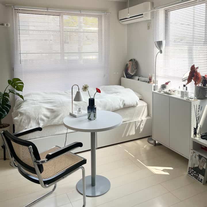 白とナチュラルトーンの寝室、ラタン椅子と小さなテーブル、フロアランプが配置された雰囲気のあるインテリア