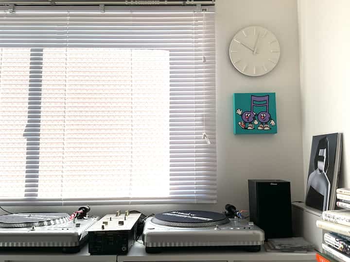 White-toned compact room featuring blinds, turntables, a wall clock, and artwork creating a minimal music workspace