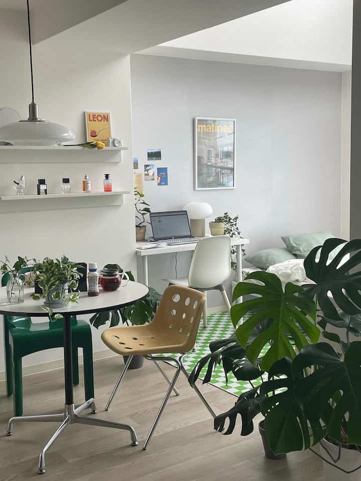 A white-themed studio apartment space with green plants, a desk, beige chair, and cafe-inspired multifunctional layout