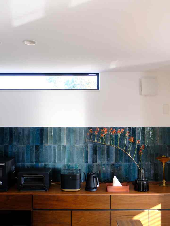 青いタイルの壁と茶色の木製キャビネットがあるキッチンスペース、黒い花瓶に赤い実の枝が飾られている印象的なインテリア