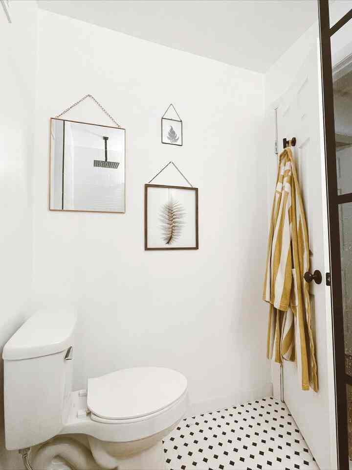 White-toned bathroom featuring a gold square mirror with chain and framed botanical prints, creating a clean unique atmosphere