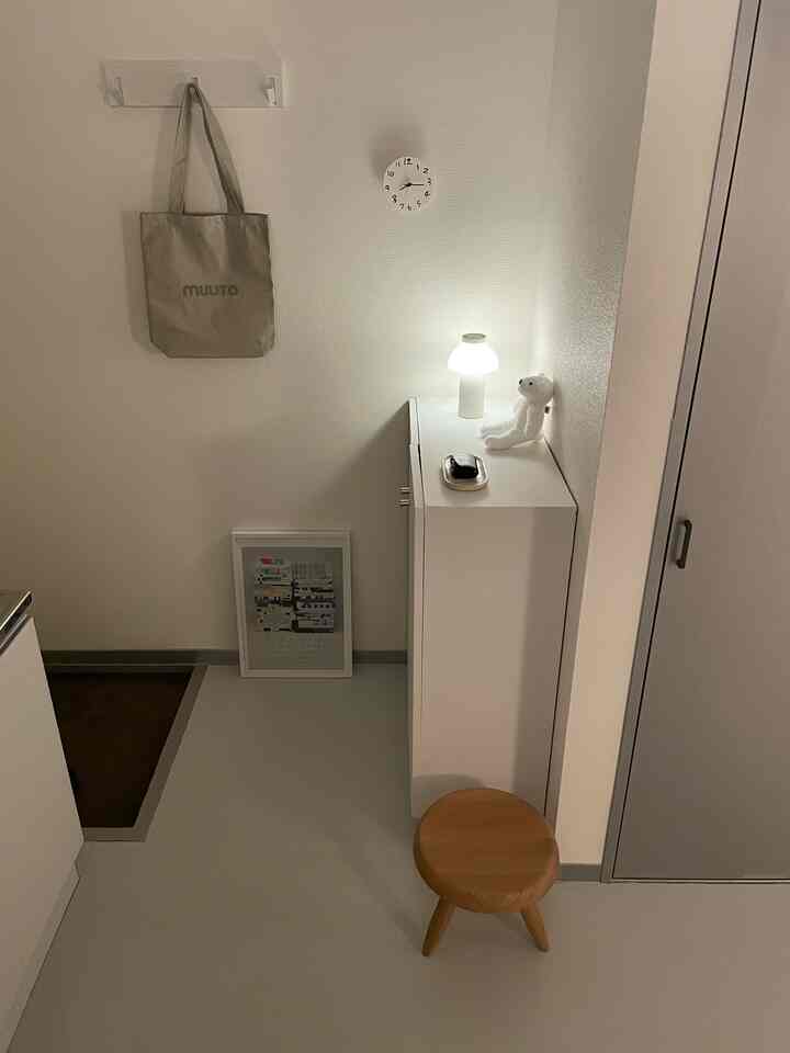 白と茶色のトーンの狭い玄関、左壁に時計とバッグ、右側に照明とぬいぐるみが置かれたキャビネットが配置された清潔感のあるインテリア