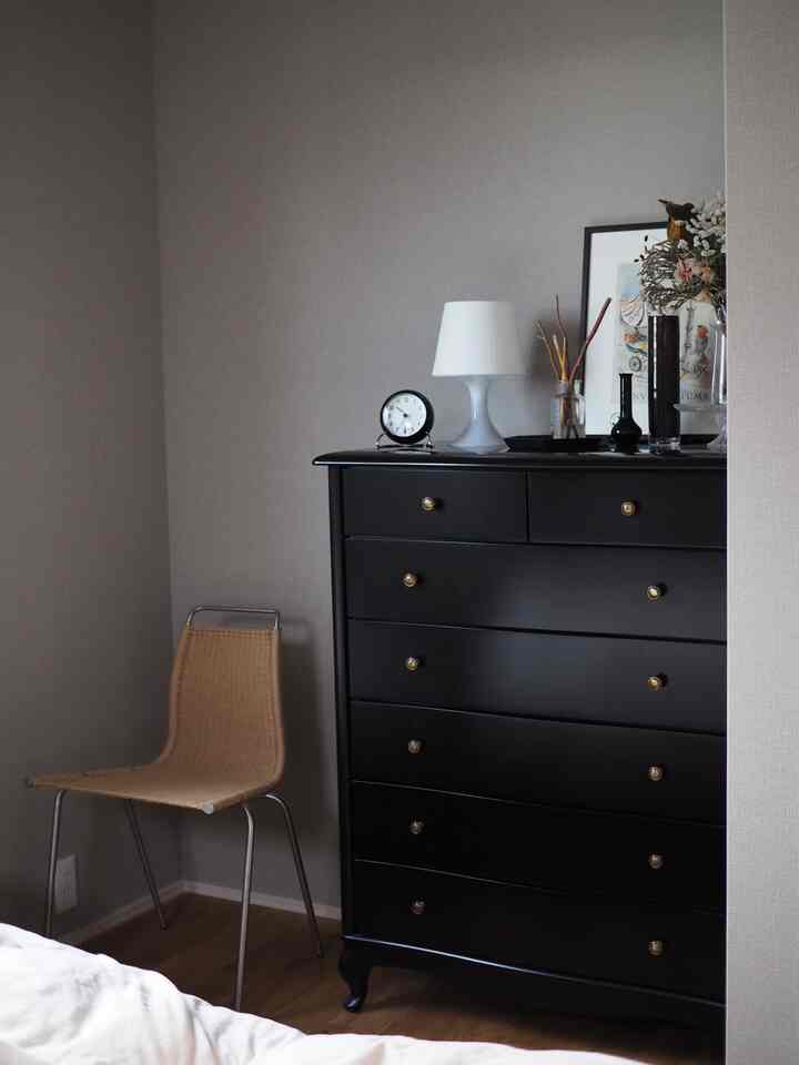 グレーの壁と黒いチェストが配置された寝室の一角、木目調の床とラタンチェアがあるミニマルなインテリア