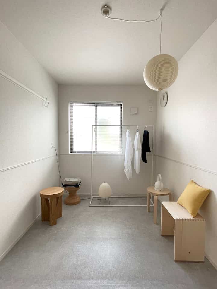 Bright white-toned small room featuring a simple iron hanger rack, natural wood furniture, and a yellow cushion as an accent